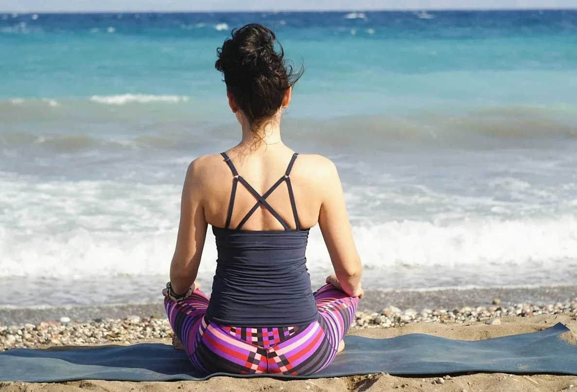 Woman sitting on a mat on a pebble beach, facing the sea
