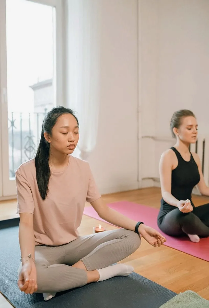 Two women sitting cross-legged with eyes closed on mats in a bright room