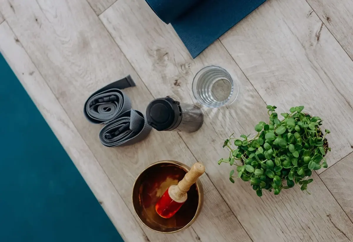 Yoga mat, strap, glass of water, singing bowl and a small potted herb on a wooden floor