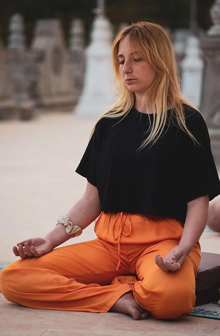Woman in a black top and orange trousers sitting cross-legged with eyes closed