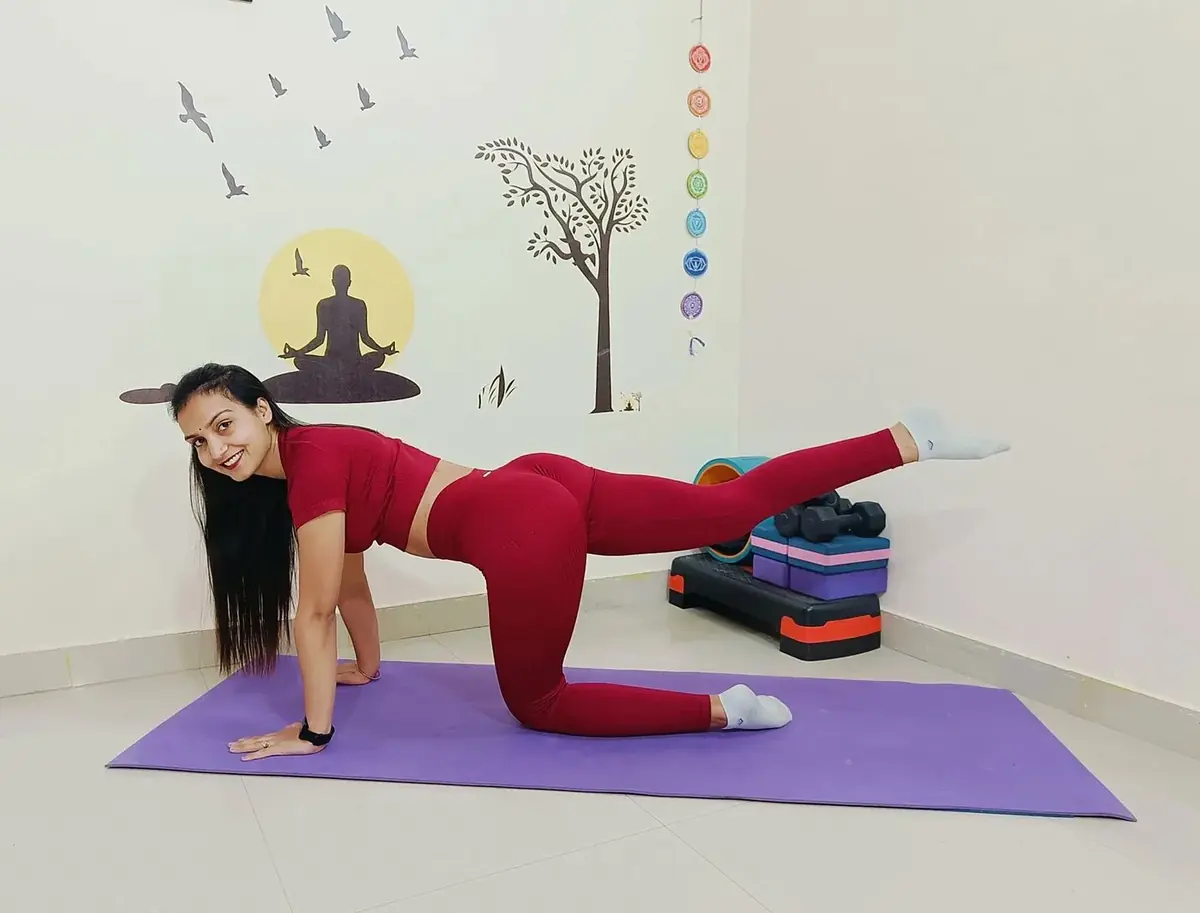 Woman in red practising a kneeling leg-lift on a purple mat in a bright room