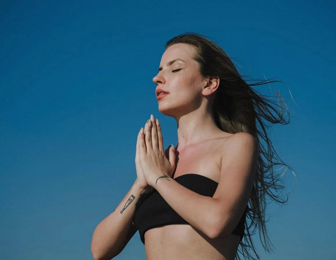 Woman with eyes closed and hands pressed together in front of her chest against a blue sky