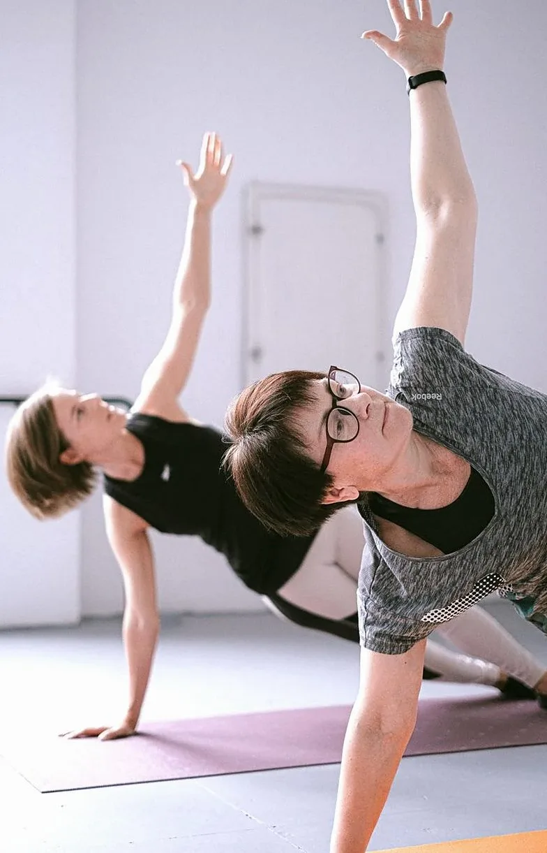Two women in a side-stretch pose with arms raised in a bright room